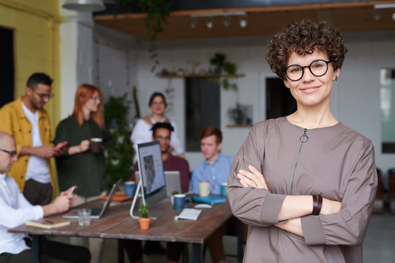 our-mission Smiling businesswoman with curly hair stands confidently in a modern office space with colleagues.